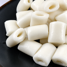 Gros plan sur des gâteaux de riz coréens blancs en forme de tubes creux, à la texture lisse et moelleuse, disposés sur un plateau noir.