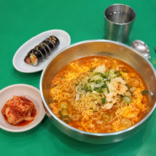 Ramen coréen épicé avec œuf et oignons verts, servi dans un bol métallique avec du kimchi et du gimbap, dans un style de repas coréen traditionnel.