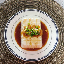 Tofu moelleux rectangulaire quadrillé, nappé de sauce soja avec ail et huile pimentée, garni d’oignons verts et de graines de sésame, présenté dans une assiette blanche à liseré sur un set de table tressé – ACEMARTmall Paris
