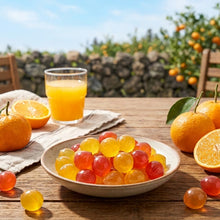 Une assiette de petites gelées rondes et translucides (jaunes et rouges) à la mandarine de Jeju, posée sur une table en bois en plein air. Autour, des mandarines fraîches, un verre de jus de fruits et, en arrière-plan, un paysage naturel ensoleillé de Jeju avec ses arbres et ses murs de pierre.