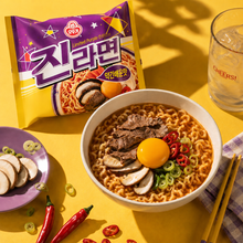Photo de mise en scène du Jin Ramen légèrement épicé Ottogi 120 g. Sur une table jaune, le sachet violet et jaune du Jin Ramen légèrement épicé est placé à l’arrière, tandis qu’un bol de ramen garni de bœuf, de champignons, d’un œuf mollet, de piments rouges et de ciboule est disposé au premier plan. Autour du bol se trouvent une assiette violette avec des champignons et de la ciboule tranchés, des piments rouges, des baguettes en bois et un verre rempli de glaçons. L’éclairage jaune chaleureux et les ombre