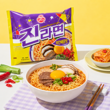 Photo de mise en scène du Jin Ramen légèrement épicé Ottogi 120 g. Le sachet violet et jaune du Jin Ramen légèrement épicé est placé à l’arrière, tandis qu’un bol de ramen garni de bœuf, de champignons, d’un jaune d’œuf, de piments rouges et de ciboule est présenté au premier plan. À côté du bol se trouvent de la ciboule et des piments rouges coupés, tandis qu’un petit bol de kimchi est visible à l’arrière-plan, à droite. Un tissu à carreaux violets est placé sous le bol et des baguettes en bois sont posées