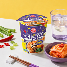 Photo de mise en scène du Jin Ramen légèrement épicé Ottogi en petit bol de 65 g, accompagné de kimchi, de ciboule, de piments rouges, d’un verre rempli de glaçons et de baguettes en bois.